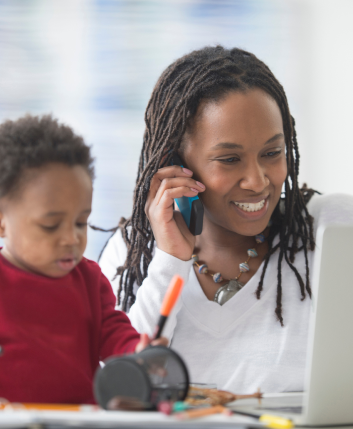 Mulher negra falando ao telefone em frente ao computador com o filho pequeno ao lado, no colo, desenhando com uma caneta colorida